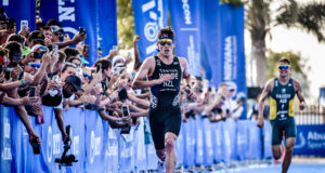 Two male triathletes race down a blue carpet lined with fans