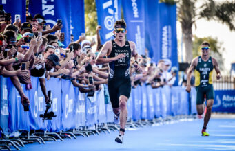 Two male triathletes race down a blue carpet lined with fans