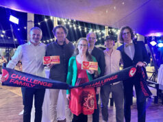 6 people (1 of them a woman) hold up a Challenge Family finish tape and certificates in a room that looks dressed for an awards ceremony.