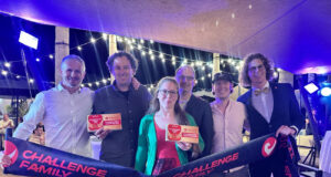 6 people (1 of them a woman) hold up a Challenge Family finish tape and certificates in a room that looks dressed for an awards ceremony.