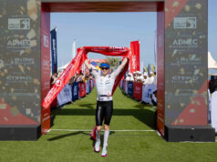 A woman crosses the finish line with red finish tape held above her head and a smile on her face