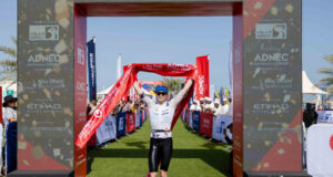 A woman crosses the finish line with red finish tape held above her head and a smile on her face