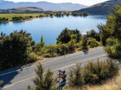 A cyclist cycles on a smooth road next to a tree lined lake