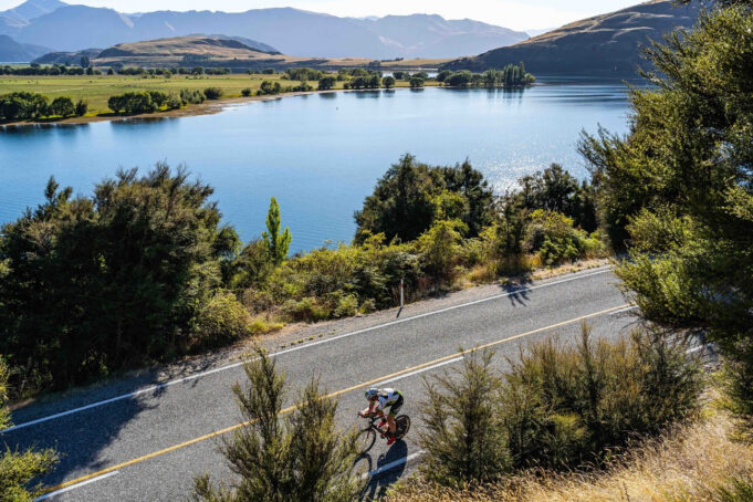 A cyclist cycles on a smooth road next to a tree lined lake