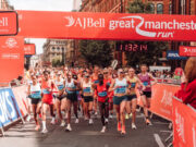 UKA 10K Road Race Champs return to Manchester Elite athletes run under a red gantry at the Great Manchester Run. The road ahead is clear, and both sides of the road are lined with spectators.