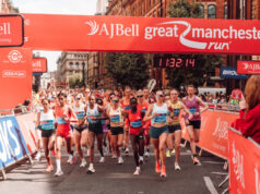 UKA 10K Road Race Champs return to Manchester Elite athletes run under a red gantry at the Great Manchester Run. The road ahead is clear, and both sides of the road are lined with spectators.