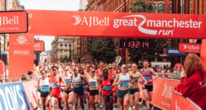 Elite athletes run under a red gantry at the Great Manchester Run. The road ahead is clear, and both sides of the road are lined with spectators.
