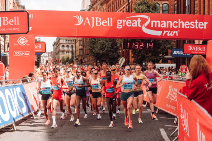Elite athletes run under a red gantry at the Great Manchester Run. The road ahead is clear, and both sides of the road are lined with spectators.