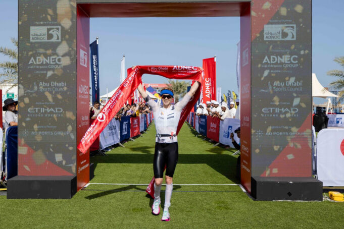 A woman crosses the finish line with red finish tape held above her head and a smile on her face
