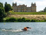 A swimmer wearing an orange swim hat swims in a lake past Blenheim Palace