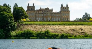 A swimmer wearing an orange swim hat swims in a lake past Blenheim Palace