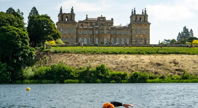 A swimmer wearing an orange swim hat swims in a lake past Blenheim Palace