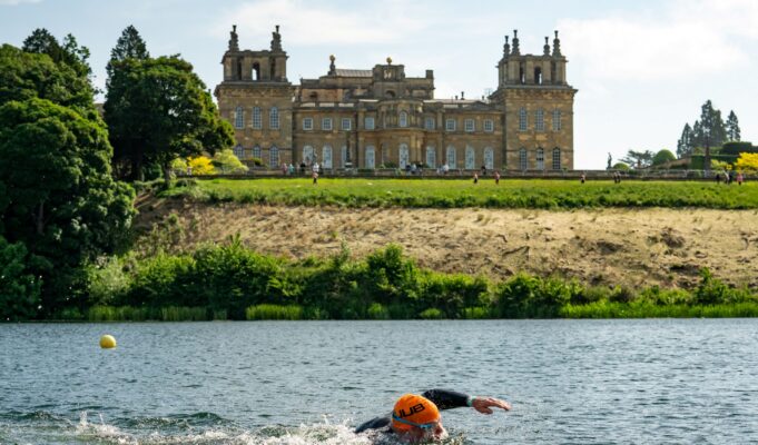 A swimmer wearing an orange swim hat swims in a lake past Blenheim Palace