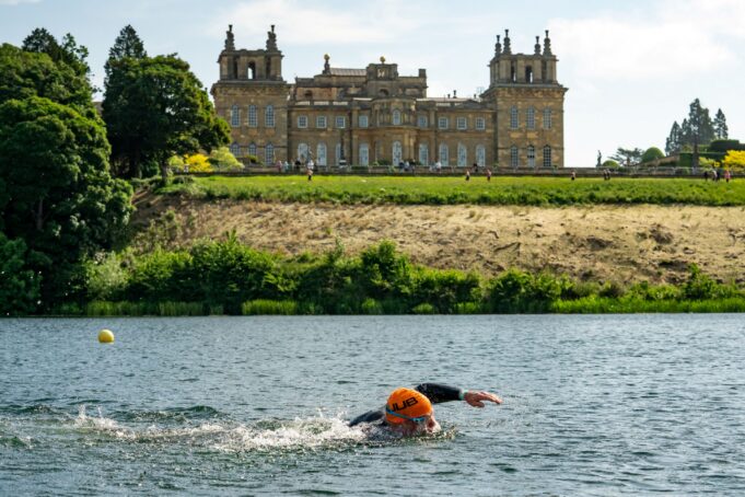 A swimmer wearing an orange swim hat swims in a lake past Blenheim Palace