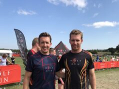 Two men in sports kit pose together on a muddy field, smiiling