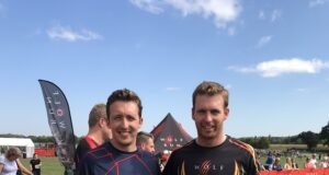 Two men in sports kit pose together on a muddy field, smiiling