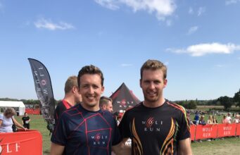 Two men in sports kit pose together on a muddy field, smiiling
