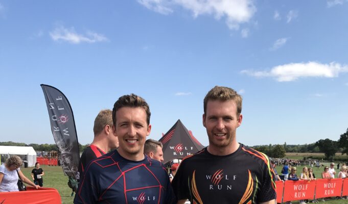 Two men in sports kit pose together on a muddy field, smiiling
