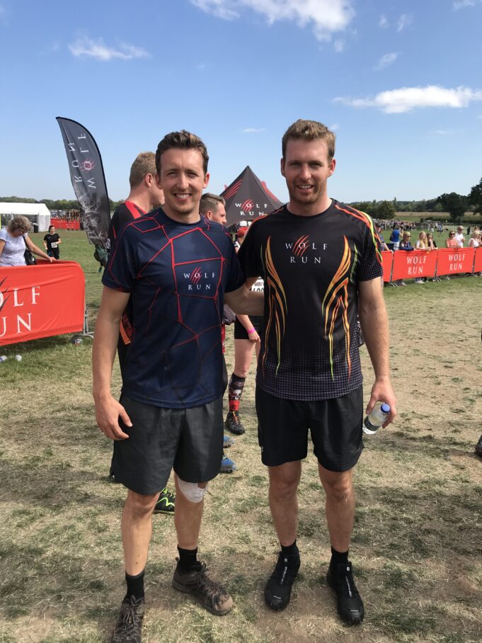 Two men in sports kit pose together on a muddy field, smiiling