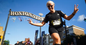 A female runner in all black swoops for the camera inn front of a tall ornate black and gold metal sign saying Hoxton Street