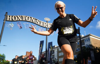 A female runner in all black swoops for the camera inn front of a tall ornate black and gold metal sign saying Hoxton Street