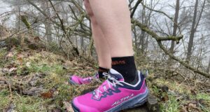 A runner wears pink shoes and black socks. Only the bottom parts of the legs and feet are visible in a grassy/rocky environment
