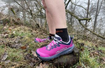 A runner wears pink shoes and black socks. Only the bottom parts of the legs and feet are visible in a grassy/rocky environment