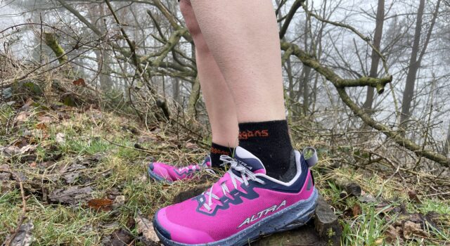 A runner wears pink shoes and black socks. Only the bottom parts of the legs and feet are visible in a grassy/rocky environment