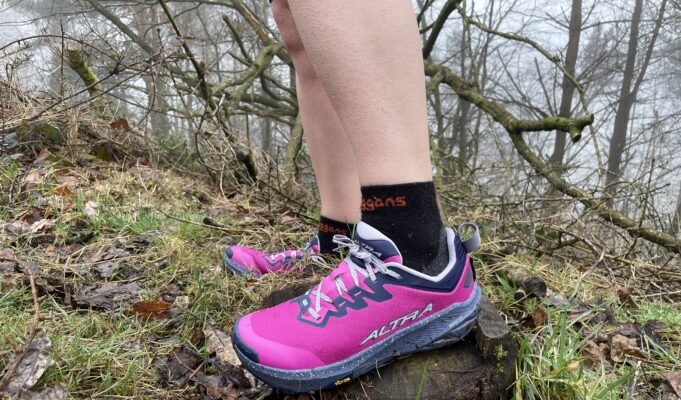 A runner wears pink shoes and black socks. Only the bottom parts of the legs and feet are visible in a grassy/rocky environment