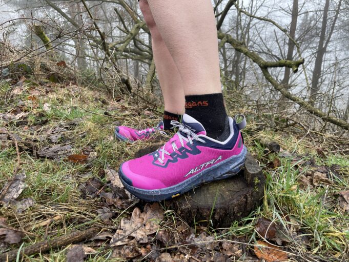 A runner wears pink shoes and black socks. Only the bottom parts of the legs and feet are visible in a grassy/rocky environment