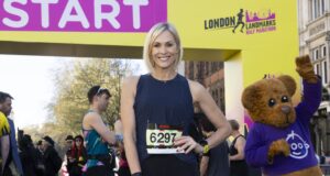 A woman with a blonde bob wearing black vest and leggings and a race number poses at the start line with a hand on her hip.