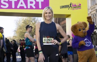 A woman with a blonde bob wearing black vest and leggings and a race number poses at the start line with a hand on her hip.