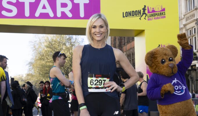 A woman with a blonde bob wearing black vest and leggings and a race number poses at the start line with a hand on her hip.