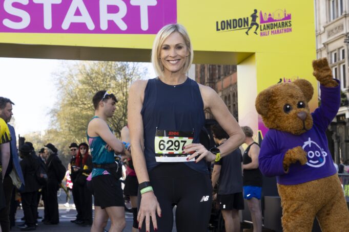 A woman with a blonde bob wearing black vest and leggings and a race number poses at the start line with a hand on her hip.
