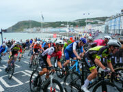 Brightly coloured road cyclists in a Welsh bay. Guest houses and the sea are visible.
