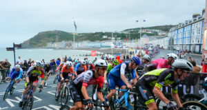 Brightly coloured road cyclists in a Welsh bay. Guest houses and the sea are visible.