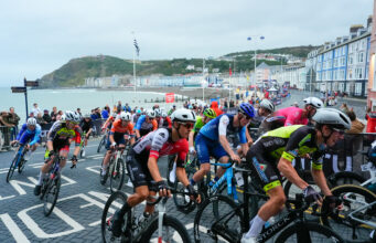 Brightly coloured road cyclists in a Welsh bay. Guest houses and the sea are visible.