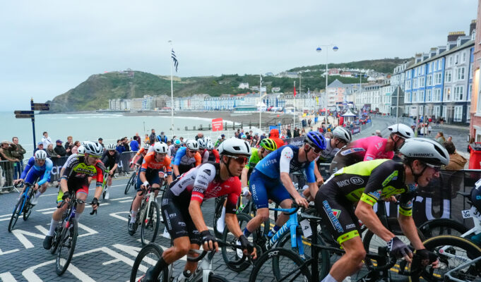 Brightly coloured road cyclists in a Welsh bay. Guest houses and the sea are visible.