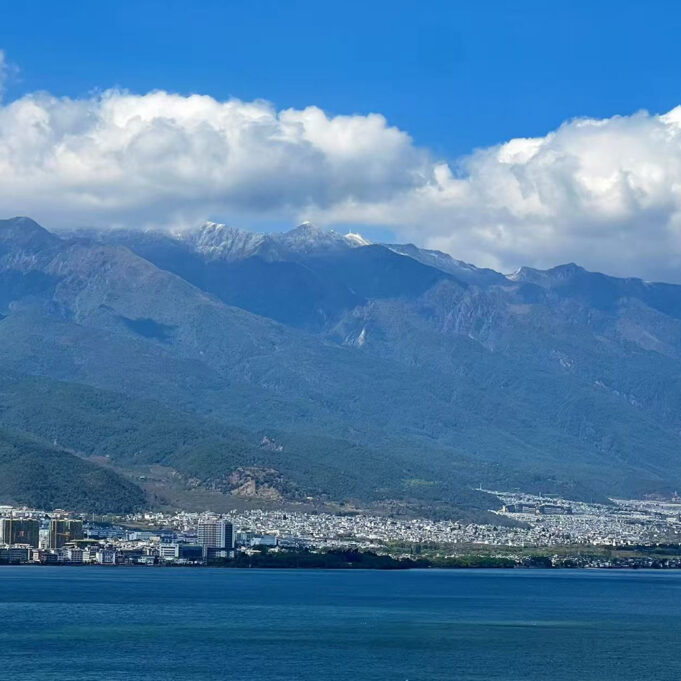 A bay of blue water with rocky mountains behind it. A city nestles between mountains and sea