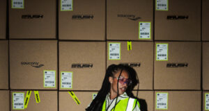 Woman stands on a pallet holding the handle of a pallet truck. She is wearing a hi vis jacket over sports kit and not looking directly at the camera.