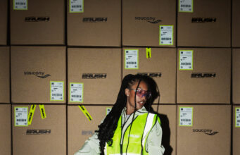 Woman stands on a pallet holding the handle of a pallet truck. She is wearing a hi vis jacket over sports kit and not looking directly at the camera.