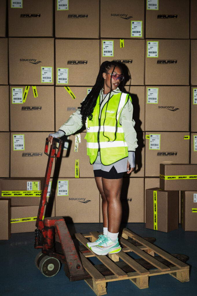 Woman stands on a pallet holding the handle of a pallet truck. She is wearing a hi vis jacket over sports kit and not looking directly at the camera.