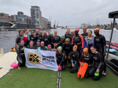 A group of women by a dock all wearing wetsuits and swimming hats pose for a phote