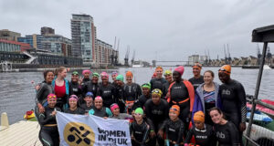 A group of women by a dock all wearing wetsuits and swimming hats pose for a phote