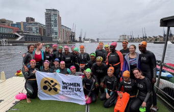 World of Triathlon LIVE Supports Tri It Forward A group of women by a dock all wearing wetsuits and swimming hats pose for a phote