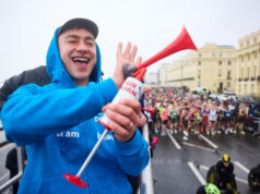 Brighton Half Marathon attracts record numbers A man wearing a blue zip up jacket and a black cap smiles as he holds an air horn. Below him, many runners on a wet start line are visible.