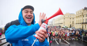 A man wearing a blue zip up jacket and a black cap smiles as he holds an air horn. Below him, many runners on a wet start line are visible.