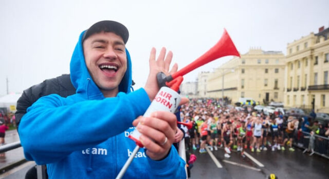 A man wearing a blue zip up jacket and a black cap smiles as he holds an air horn. Below him, many runners on a wet start line are visible.