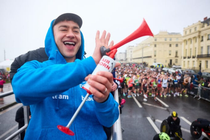 A man wearing a blue zip up jacket and a black cap smiles as he holds an air horn. Below him, many runners on a wet start line are visible.