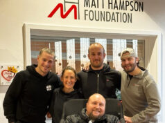 5 people pose for the camera. 4 people stand wearing running kit behind a man in a wheelchair in a coat and a blanket over his legs